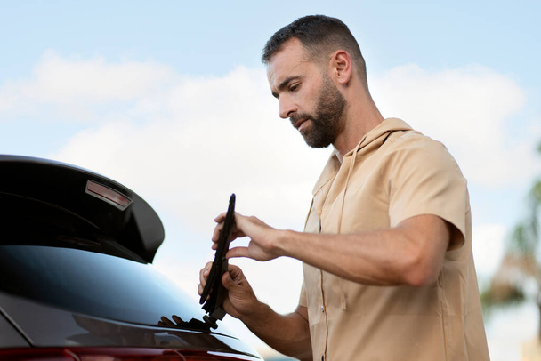 Pensive car service worker replace windshield wipers on car standing outdoors - Photo, Image