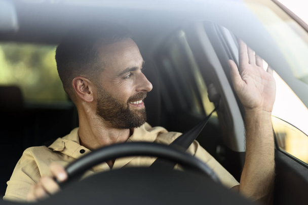 Zelfverzekerde lachende taxichauffeur zwaaiende hand wachtend op cliënt zittend in auto. Transportconcept - Foto, afbeelding