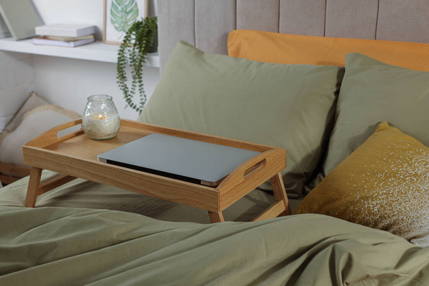 Wooden tray with modern laptop and burning candle on bed indoors - Valokuva, kuva