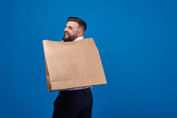 A man with a large brown paper bag on his back stands on a blue background in the studio. Package for close-up purchases. Space for a logo on a paper bag. - Zdjęcie, obraz
