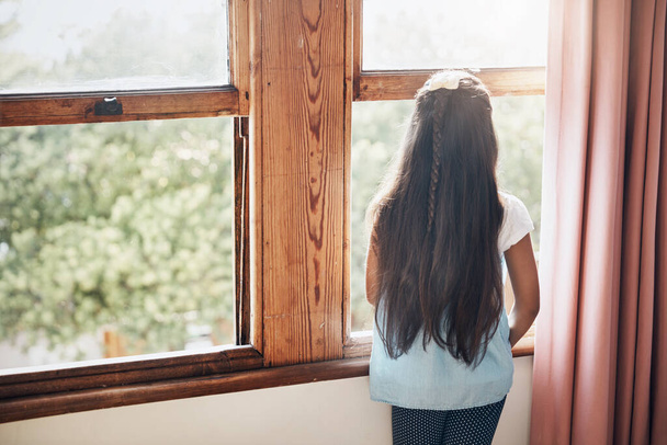 What waits for her in a great big world. Rearview shot of a young girl looking out of the window at home - Foto, Bild