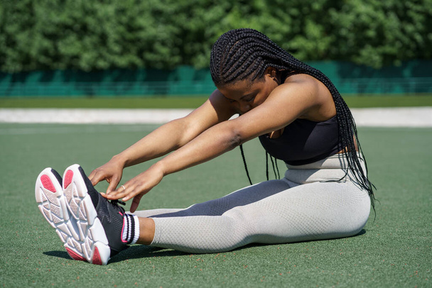 Black fit young woman in sportswear tries to reach toes with hands sitting on green turf flooring on sports ground against trees in city park. African American female enjoys stretching on sunny day - Фото, зображення