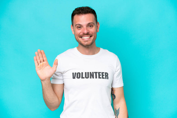 Young volunteer caucasian man isolated on blue background saluting with hand with happy expression - Foto, afbeelding