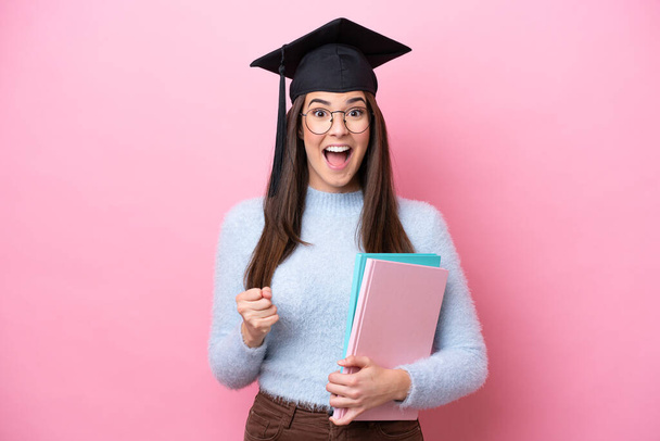 Young student Brazilian woman wearing graduated hat isolated on pink background celebrating a victory in winner position - Valokuva, kuva