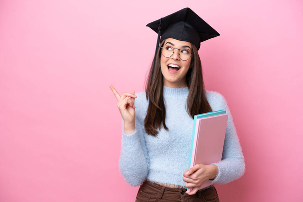 若い学生ブラジル人女性を身に着けている卒業帽子ピンクの背景に孤立指を持ち上げながら解決策を実現しようとしています - 写真・画像