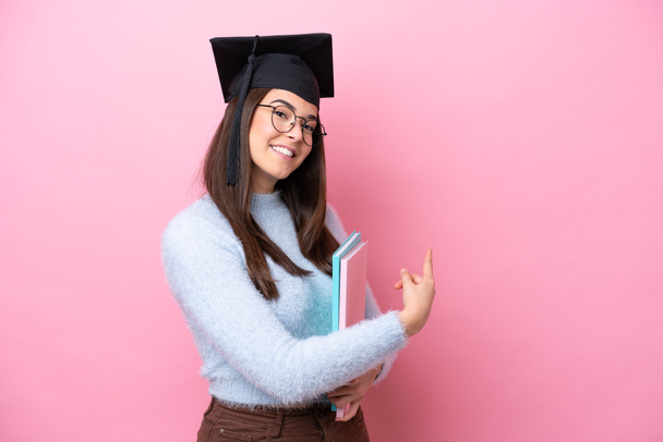 Молодая студентка бразильская женщина в шляпе выпускника изолированы на розовом фоне указывая назад - Фото, изображение