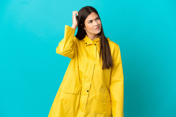 Teenager Brazilian girl wearing a rainproof coat over isolated blue background having doubts and with confuse face expression - Photo, Image