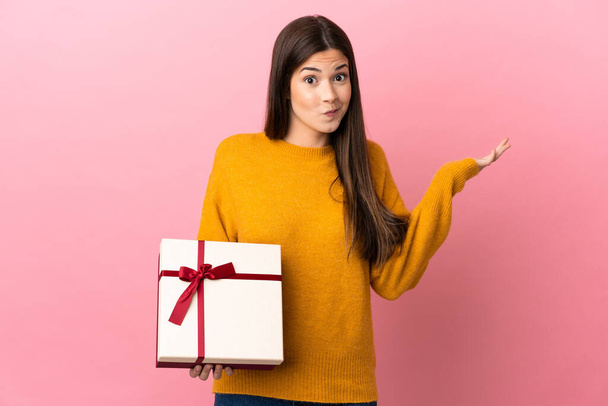 Teenager Brazilian girl holding a gift over isolated pink background having doubts while raising hands - 写真・画像