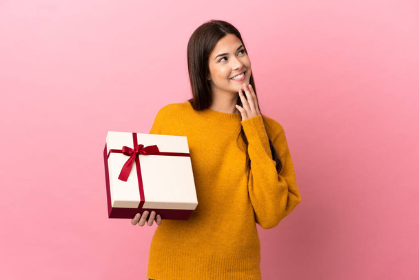 Teenager Brazilian girl holding a gift over isolated pink background looking up while smiling - 写真・画像