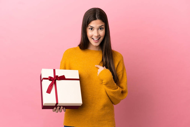 Teenager Brazilian girl holding a gift over isolated pink background with surprise facial expression - Photo, Image