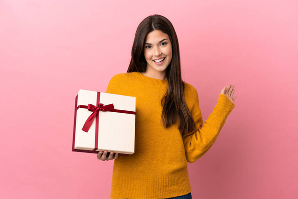 Teenager Brazilian girl holding a gift over isolated pink background saluting with hand with happy expression - Фото, изображение