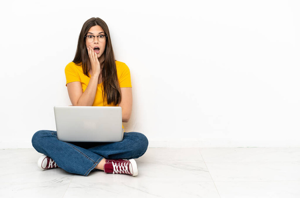 Молода жінка з ноутбуком сидить на підлозі здивована і шокована, дивлячись праворуч
 - Фото, зображення