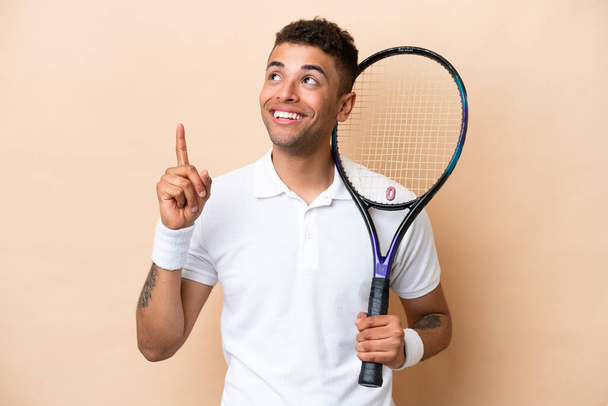 Молодой бразильский красавчик играет в теннис на бежевом фоне, указывая на отличную идею. - Фото, изображение