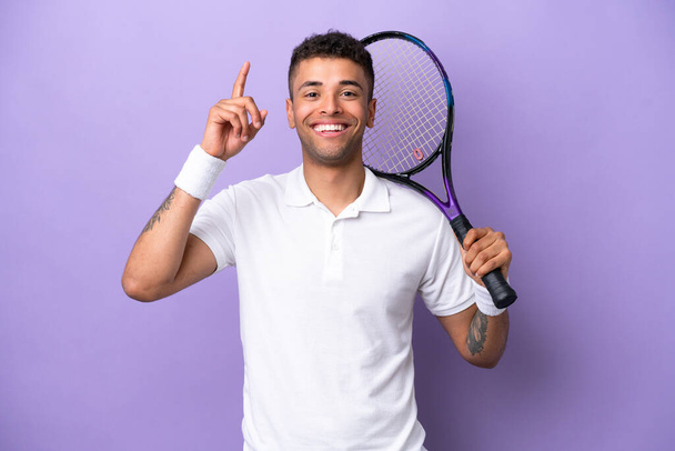 Молодой бразилец изолирован на фиолетовом фоне, играет в теннис и указывает вверх - Фото, изображение