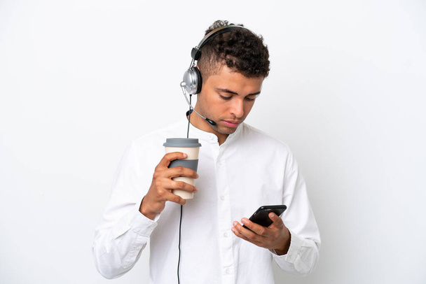 Hombre brasileño telemarketer trabajando con un auricular aislado sobre fondo blanco sosteniendo café para llevar y un móvil - Foto, imagen