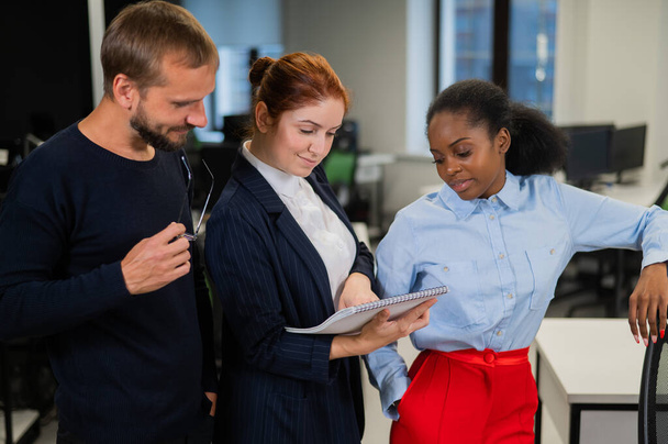 Colleagues discuss work. African young woman, caucasian man and red-haired caucasian woman communicate in the office - 写真・画像