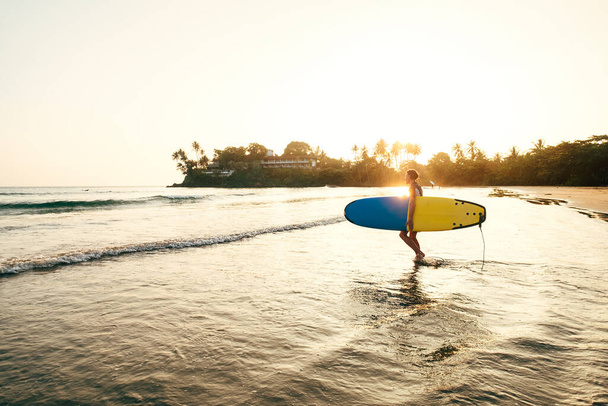 Teen boy with blue and yellow surfboard entering the waves for surfing with sunset rays on palm trees grove. Happy childhood and active sporty people vacation time concept. - Valokuva, kuva