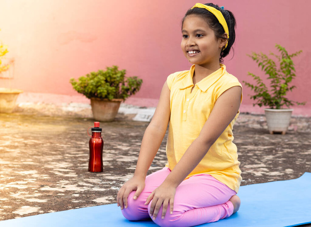 An Indian girl child practicing yoga in smiling face on yoga mat outdoors - Valokuva, kuva