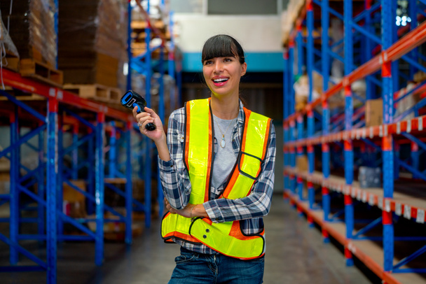 Pretty warehouse worker woman hold barcode scaner and smiling stand in the way between shelves in  workplace area. She look happy during work to delivery product to customer. - Foto, immagini