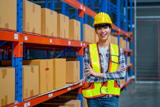 Pretty warehouse worker stand with confidence action and arm-crossed in front of shelves of product carton in workplace. - Foto, afbeelding