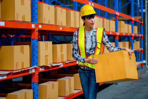 Warehouse worker woman hold the box and walk on way between shelves in workplace. Concept of professional factory worker support for delivery and transport system. - Foto, Bild