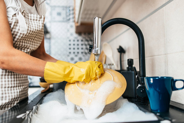 Young adult woman with yellow protective gloves washing her dishes on kitchen sink. Household and home hygiene routine. - Foto, imagen