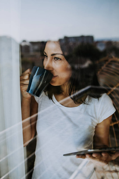 Smiled and positive woman looks out the window of her apartment or office. She is holding cup of coffee and tablet. Quarantine and social distancing during Covid-19 pandemic concept. Stay positive. - Photo, image