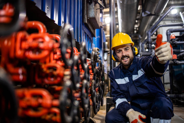 Portrait of cheerful heating plant worker standing by pipe valves and holding thumbs up. - Foto, Imagen