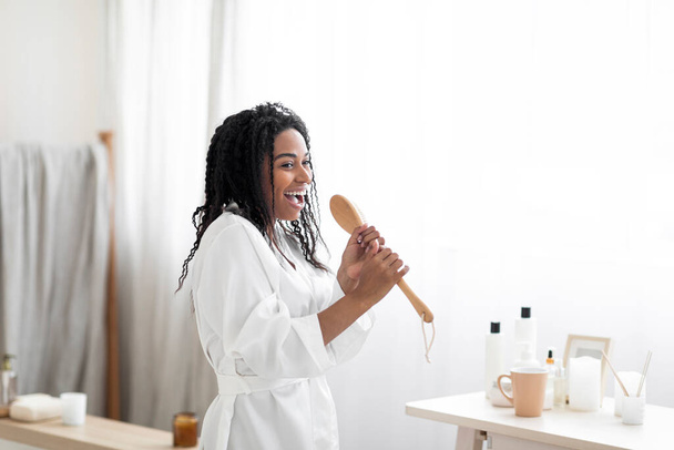 Morning Fun. Joyful African American Female Singing In Bathroom Interior, Cheerful Young Black Woman In White Silk Robe Using Body Brush As Microphone, Enjoying Home Beauty Routine, Copy Space - Foto, imagen