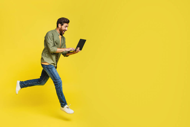 Полная длина возбужденного мужчины средних лет, использующего ноутбук и прыгающего на желтом фоне копировального пространства. Стильный мужчина-фрилансер, летающий в воздухе с современным портативным компьютером - Фото, изображение