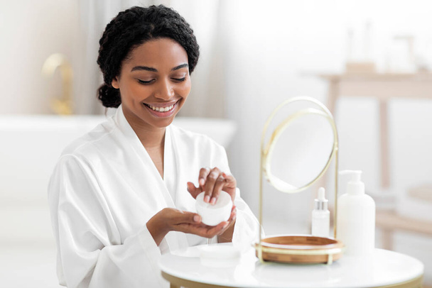 Skin Care Concept. Beautiful Black Female Using New Cosmetic At Home, Attractive African American Woman In White Silk Robe Sitting In Front Of Mirror And Applying Moisturising Face Cream, Copy Space - Valokuva, kuva