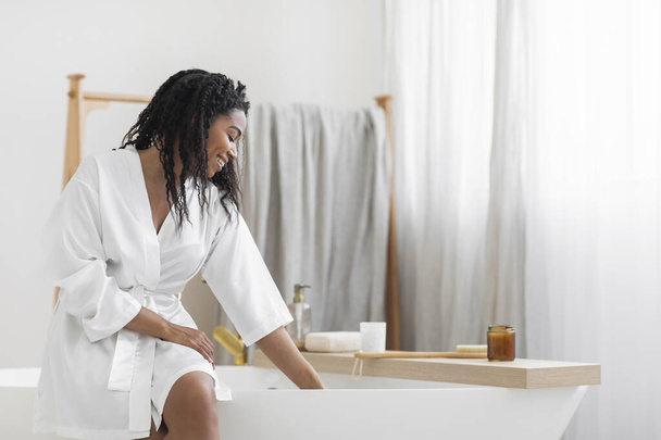 Bath Time. Beautiful Young Black Lady In White Silk Robe Sitting On Bathtub And Checking Water Temperature, Smiling African American Woman Relaxing In Modern Bathroom Interior At Home, Copy Space - Zdjęcie, obraz