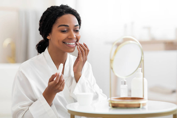 Attractive Black Female Looking At Mirror And Applying Moisturising Lip Balm, Beautiful African American Woman Holding Chapstick, Using Hygienical Lipstick While Sitting At Dressing Table At Home - Foto, afbeelding