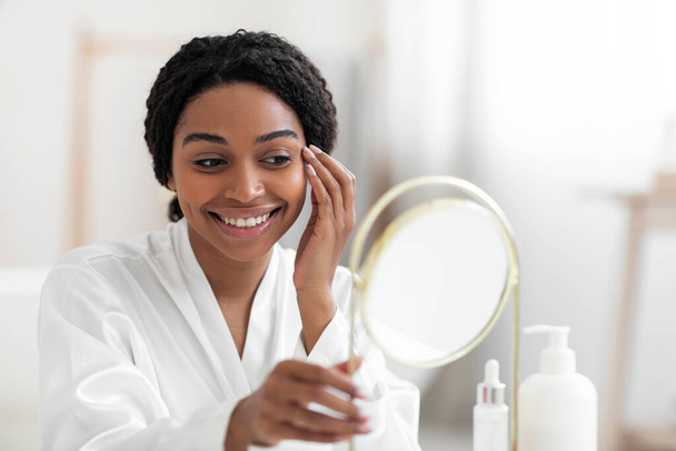 Концепція скептики. Дивовижна чорна жінка, яка дивиться на своє відображення у дзеркалі, поки робить розгул краси вдома, усміхнена афро-американська жінка в білій шовковій сукні втішає її погляд, роздягаючись - Фото, зображення