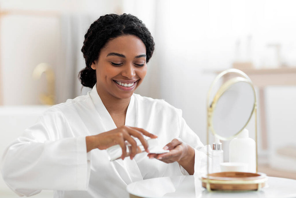 Улыбающаяся чёрная женщина заливает мицеллярную воду на хлопчатобумажную площадку, делая дома программу красоты, привлекательная молодая афроамериканская женщина сидит рядом с зеркалом, очищает лицо от макияжа, крупным планом - Фото, изображение