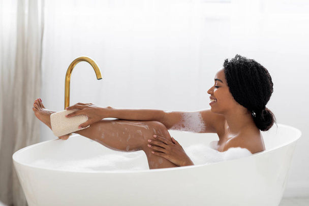 Beautiful Black Woman Taking Foamy Bath And Using Body Sponge, Happy Young African American Female Relaxing In Stylish Bathtub At Home, Enjoying Selfcare Beauty Treatments, Side View, Copy Space - 写真・画像