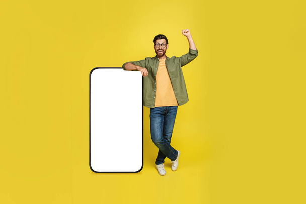 Excited man leaning on huge cellphone with blank screen and shaking fist in joy, yellow studio background. Great mobile offer concept. Mockup, full length - Photo, image