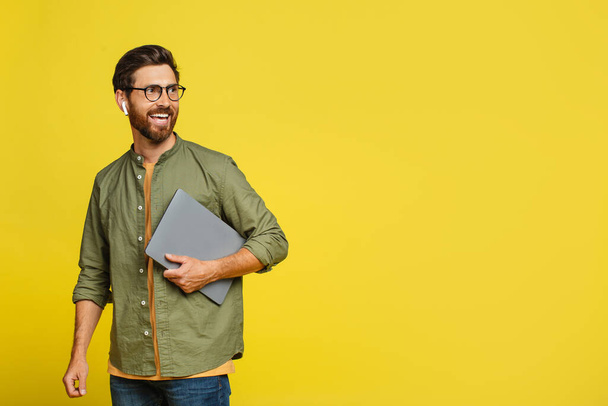 Positive stylish man in casual outfit freelancer posing with laptop over yellow studio background, looking aside at free space and smiling. Remote job concept - Foto, Imagen