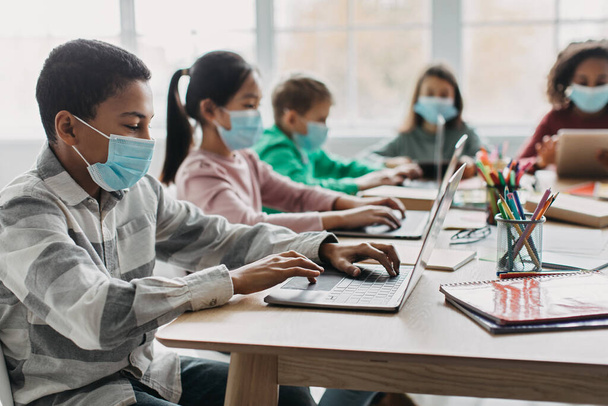 Online Education. Black Schoolboy Using Laptop Having Class With Multiracial Classmates Sitting At Desk In Modern Classroom Indoor, Wearing Face Masks. School And E-Learning. Selective Focus - Foto, imagen