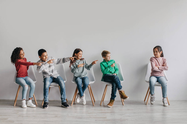 Bullying Problem At School. Mixed Kids Laughing Pointing At Victimized Asian Schoolgirl Sitting Over Gray Wall Indoors. Children Education And Social Problems Concept - Photo, Image