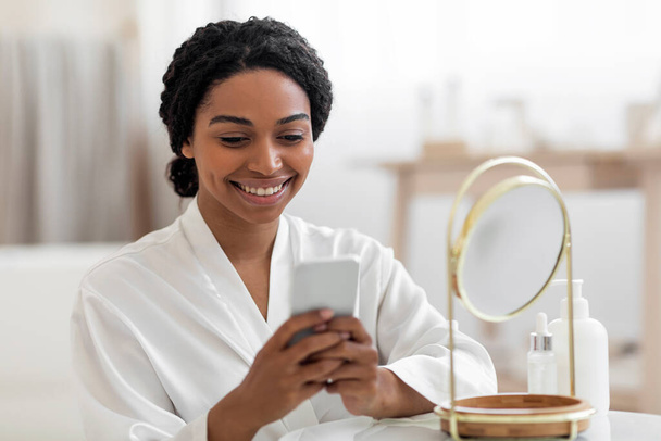 Smiling Black Woman With Smartphone Reading Beauty Hacks While Getting Ready Near Mirror At Home, Attractive African American Female Wearing White Silk Robe Browsing Internet On Mobile Phone - 写真・画像