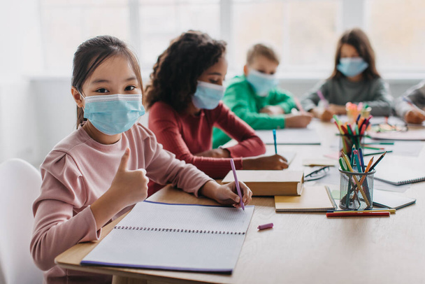 I Like My School. Asian Kid Girl Gesturing Thumbs Up Wearing Face Mask Approving Modern Education Sitting At Desk With Multiethnic Classmates In Classroom Indoors. Selective Focus - Photo, image