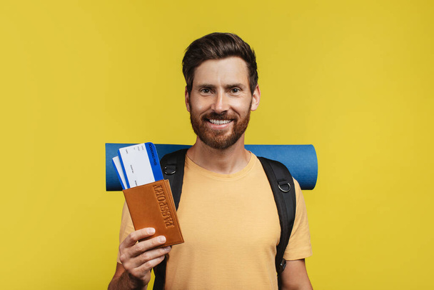 Middle aged male traveler with rucksack holding international passport and airplane tickets over yellow studio background. Happy man ready to go on vacation abroad - Foto, Imagen