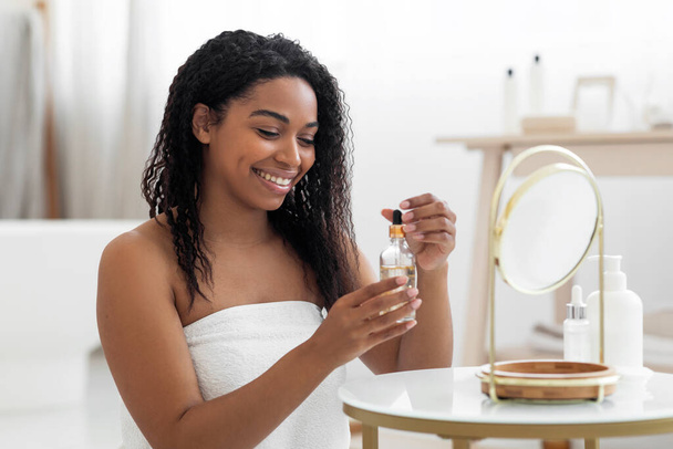 Happy Black Lady Holding Bottle With Moisturising Serum For Skincare, Beautiful African American Woman Wrapped In Bath Towel Sitting At Dressing Table, Making Beauty Routine At Home, Closeup - Foto, imagen