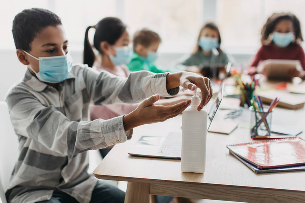 Coronavirus Protection. Black Schoolboy Disinfecting Hands With Sanitizer Wearing Protective Face Mask Sitting With Diverse Classmates Kids In Modern Classroom. School And Healthcare - 写真・画像