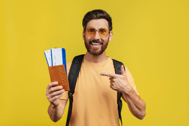 Positive middle aged man with backpack pointing at passport and airplane tickets, posing over yellow studio background and smiling at camera - Zdjęcie, obraz