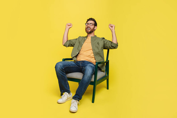 Emotional middle aged man in casual raising fists up, sitting in armchair on yellow studio background, celebrating success, happy male sharing good emotions, copy space - Photo, Image