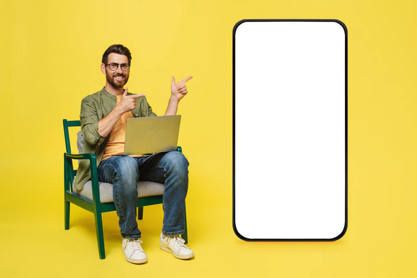 Excited middle aged man sitting on chair using laptop pc and pointing at big cellphone with empty white screen, presenting new cool app, copy space mockup, website design banner - Fotografie, Obrázek