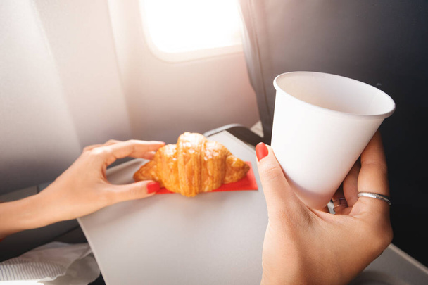 A girl passenger eats a croissant and drinks coffee sitting by the window on an airplane and flies on vacation - Φωτογραφία, εικόνα