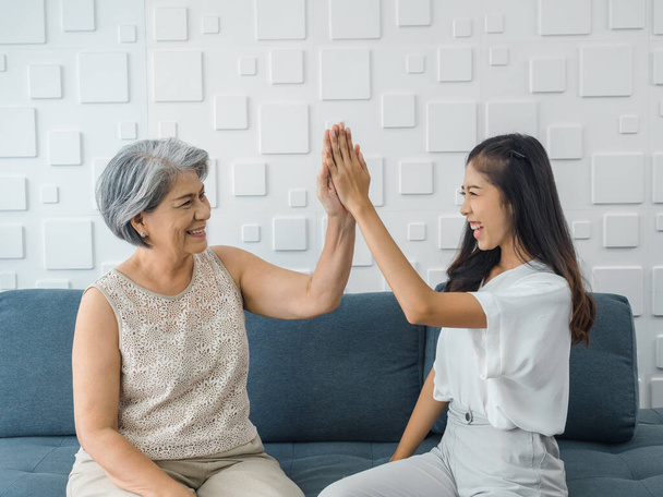 Asian young woman, cheerful daughter giving high five to her mom, old senior female, sitting on grey sofa seat in white room. Mother and daughter celebrating victory win together, clapping hands. - Foto, imagen